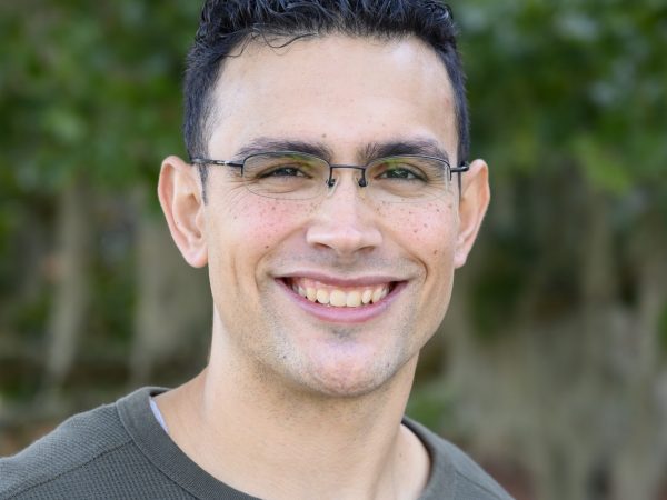 Person smiles at camera while standing in front of trees outside.